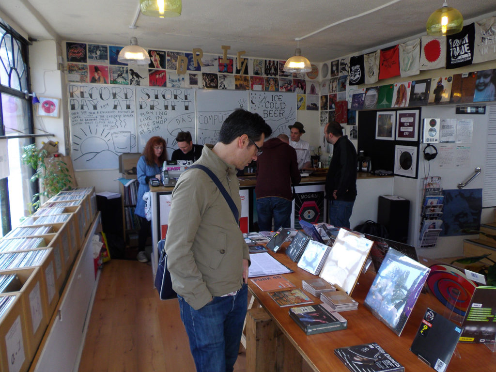 Customers browsing and buying records in a record store. Records are on display on a central table and in rows of boxes, while the walls are decorated with album covers, t-shirts, and a sign about Record Store Day.